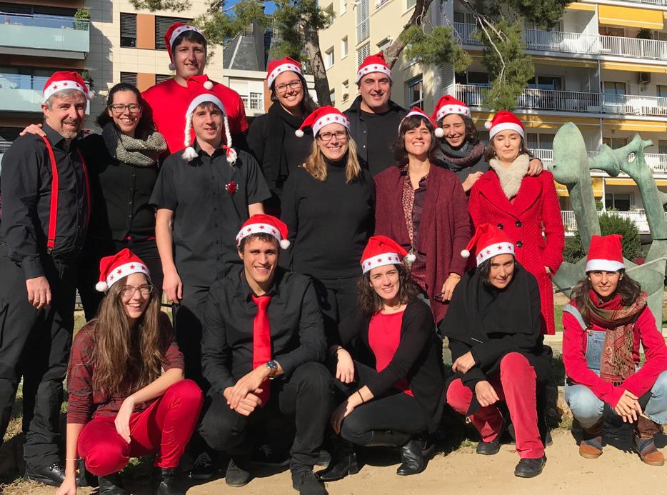 Els cantaires d'En Clau Vocal, amb barrets de Pare Noel i roba negra i vermella, fan una foto de grup al carrer en un dia assolellat.