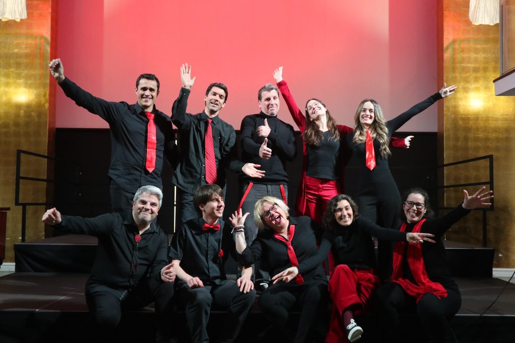 Els cantaires d'En Clau Vocal, de negre amb corbates vermelles, posen amb els braços oberts davant d'un fons de llum vermella a l'escenari del Centre Cívic Pere Pruna.