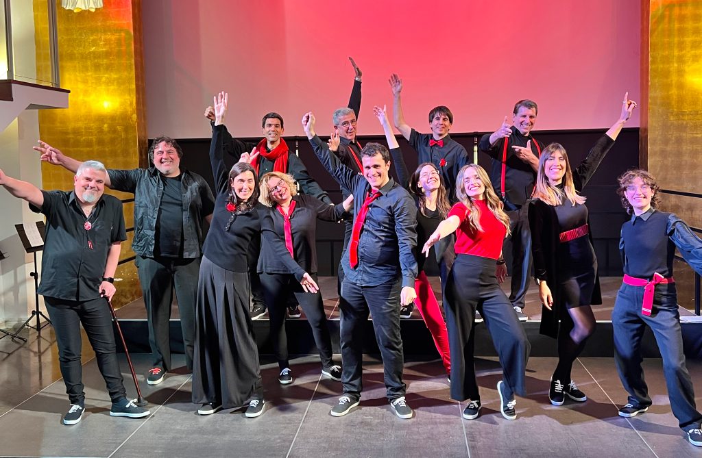 Els cantaires d'En Clau Vocal saluden amb els braços enlaire a l'escenari del Centre Cívic Pere Pruna, il·luminat amb llum vermella.